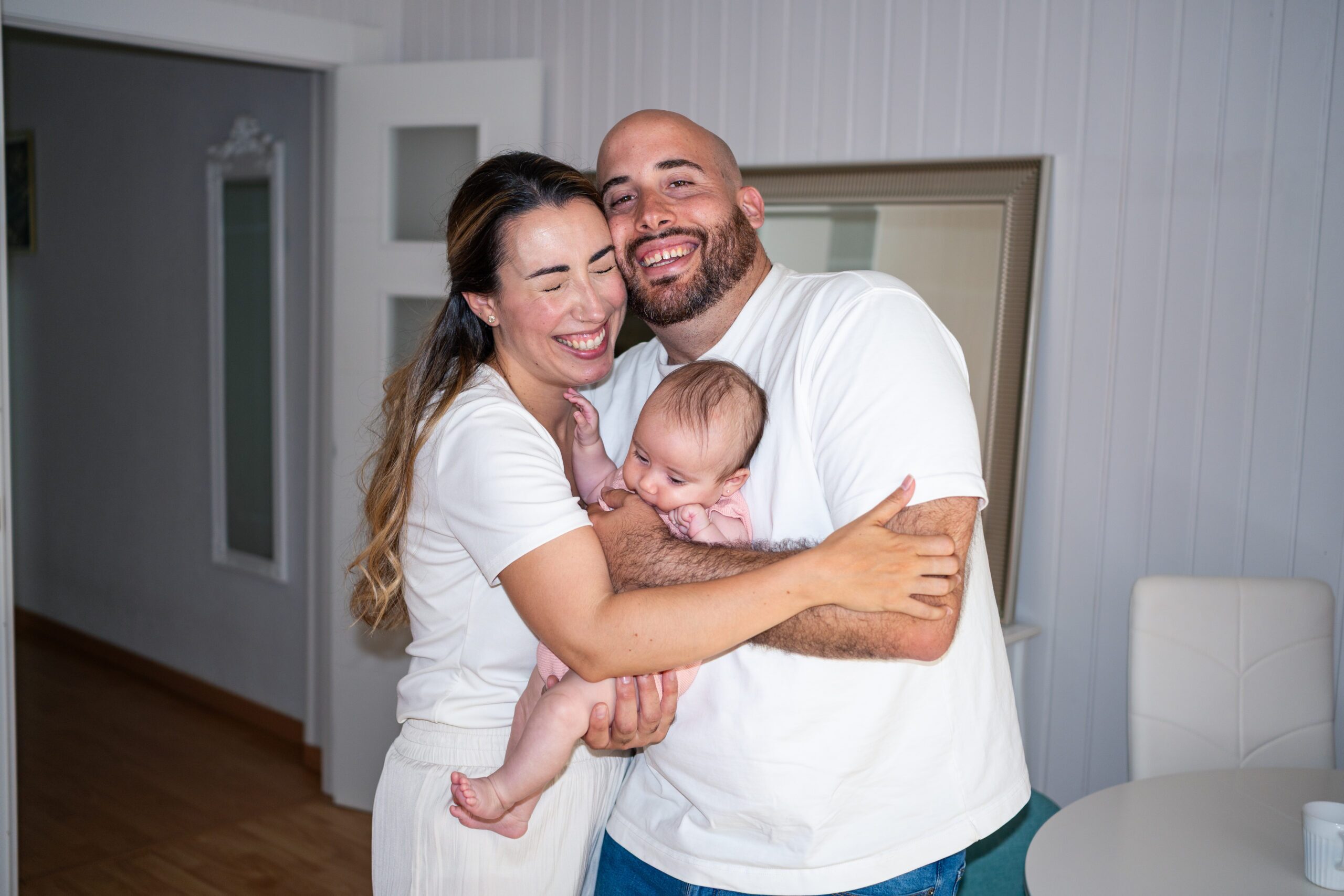 A joyful family shares a heartfelt moment in their cozy home, embracing their baby with smiles. Candid shot captured with direct flash, enhancing their loving connection and happiness together.